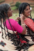 Two women are sitting outdoors, engaging in the activity of braiding hair. They are wearing traditional garments with intricate patterns, and one woman is smiling while braiding the other's hair.