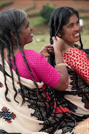 Two women are sitting outdoors, engaging in the activity of braiding hair. They are wearing traditional garments with intricate patterns, and one woman is smiling while braiding the other's hair.