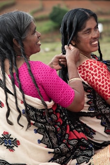 Two women are sitting outdoors, engaging in the activity of braiding hair. They are wearing traditional garments with intricate patterns, and one woman is smiling while braiding the other's hair.
