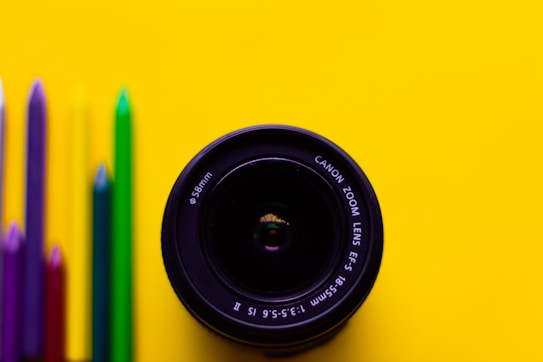 A camera lens is positioned on a vibrant yellow background. To the left of the lens, a row of colored pencils is neatly aligned, featuring hues of purple, red, green, and blue.