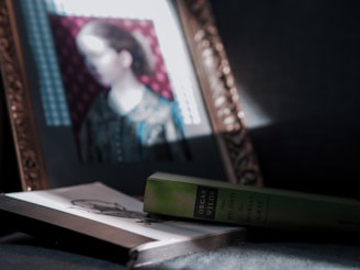 A framed portrait of a woman, softly lit from the side, is positioned above a closed book titled 'Das Bildnis des Dorian Gray' by Oscar Wilde. The scene is set against a dark background, creating a dramatic play of light and shadow.