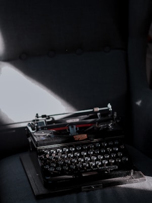 An old typewriter sits on a dark, upholstered chair. Light streams in from the left, creating a soft highlight on the black surface of the typewriter and casting shadows across its keys. The scene is muted and antique, evoking a sense of nostalgia.