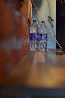 Two clear plastic water bottles with blue labels are placed side by side on a wooden shelf. The background features a red brick wall on the left and a light blue wall on the right, with a metal chain hanging down. Some wires are visible in the top right corner.