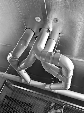 Several metallic pipes with multiple bends and turns are attached to a ceiling. The surface of the ceiling appears textured, and a chain-link fence is visible in the lower part of the image.