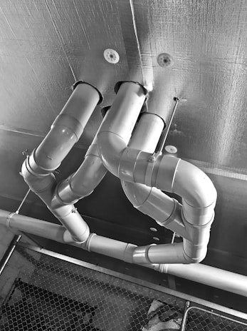 Several metallic pipes with multiple bends and turns are attached to a ceiling. The surface of the ceiling appears textured, and a chain-link fence is visible in the lower part of the image.