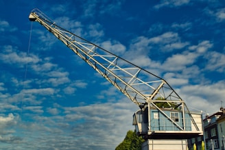 A large, metal crane stands prominently against a bright blue sky dotted with white clouds. The crane's structure is made of a metallic lattice with a cab at one end, perched on a building set amidst greenery. The design is industrial, with geometric precision providing a stark contrast to the natural elements visible at the horizon.