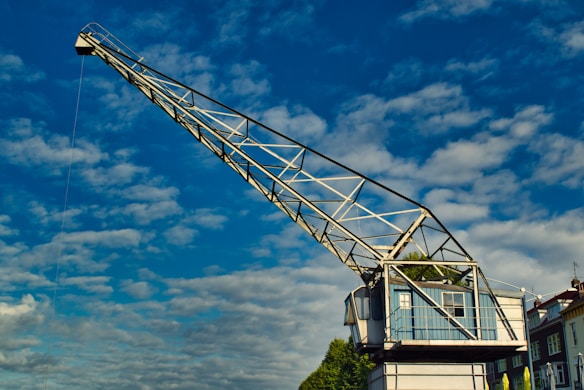 A large, metal crane stands prominently against a bright blue sky dotted with white clouds. The crane's structure is made of a metallic lattice with a cab at one end, perched on a building set amidst greenery. The design is industrial, with geometric precision providing a stark contrast to the natural elements visible at the horizon.