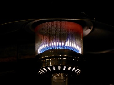 A close-up view of a metallic outdoor heater with a visible flame inside a perforated mesh enclosure. The heater radiates a warm glow with colors ranging from blue to yellow, surrounded by a dark background.