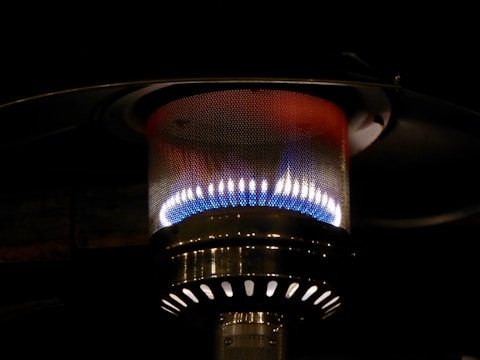 A close-up view of a metallic outdoor heater with a visible flame inside a perforated mesh enclosure. The heater radiates a warm glow with colors ranging from blue to yellow, surrounded by a dark background.