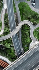 A top-down aerial view of intersecting roads and greenery. The image includes a winding pedestrian pathway and multiple roads with lanes marked by yellow lines. There is lush green vegetation, small trees, and patches of grass between the pathways. Some cars are visible in a small parking area at the top right corner.