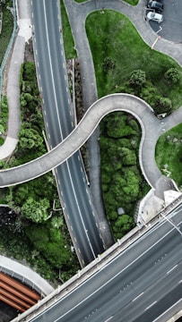 A top-down aerial view of intersecting roads and greenery. The image includes a winding pedestrian pathway and multiple roads with lanes marked by yellow lines. There is lush green vegetation, small trees, and patches of grass between the pathways. Some cars are visible in a small parking area at the top right corner.