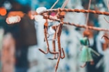 A close-up of handmade stick figures hanging from a woven branch, with blurred bokeh lights in the background. There is a figure wrapped in twine and another in a green dress.