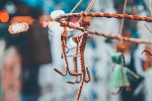 A close-up of handmade stick figures hanging from a woven branch, with blurred bokeh lights in the background. There is a figure wrapped in twine and another in a green dress.