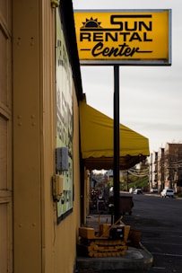 A rental business with a prominent yellow sign displaying the name 'Sun Rental Center' is featured. The building’s exterior wall is painted yellow, matching the sign and awning. There's a small piece of heavy machinery parked near the building, and a main road extends into the distance on the right side of the image with a few vehicles.