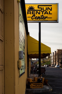 A rental business with a prominent yellow sign displaying the name 'Sun Rental Center' is featured. The building’s exterior wall is painted yellow, matching the sign and awning. There's a small piece of heavy machinery parked near the building, and a main road extends into the distance on the right side of the image with a few vehicles.