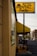 A rental business with a prominent yellow sign displaying the name 'Sun Rental Center' is featured. The building’s exterior wall is painted yellow, matching the sign and awning. There's a small piece of heavy machinery parked near the building, and a main road extends into the distance on the right side of the image with a few vehicles.