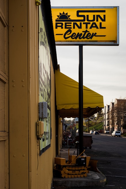 A rental business with a prominent yellow sign displaying the name 'Sun Rental Center' is featured. The building’s exterior wall is painted yellow, matching the sign and awning. There's a small piece of heavy machinery parked near the building, and a main road extends into the distance on the right side of the image with a few vehicles.