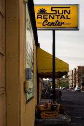 A rental business with a prominent yellow sign displaying the name 'Sun Rental Center' is featured. The building’s exterior wall is painted yellow, matching the sign and awning. There's a small piece of heavy machinery parked near the building, and a main road extends into the distance on the right side of the image with a few vehicles.