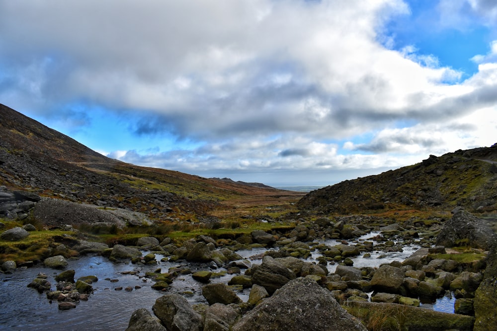 gray rocks on river near green mountain under blue and white sunny cloudy sky during daytime