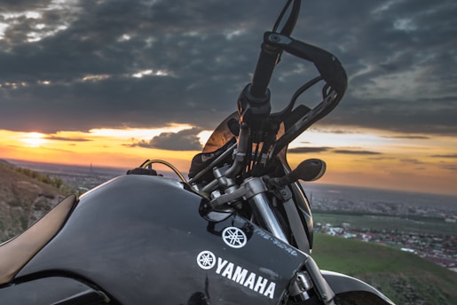 A close-up view of a Yamaha motorcycle with its handle and a part of the chassis visible against a backdrop of a picturesque sunset. The horizon shows a landscape that includes hills and a distant town or city under a sky filled with dramatic clouds.