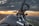 A close-up view of a Yamaha motorcycle with its handle and a part of the chassis visible against a backdrop of a picturesque sunset. The horizon shows a landscape that includes hills and a distant town or city under a sky filled with dramatic clouds.