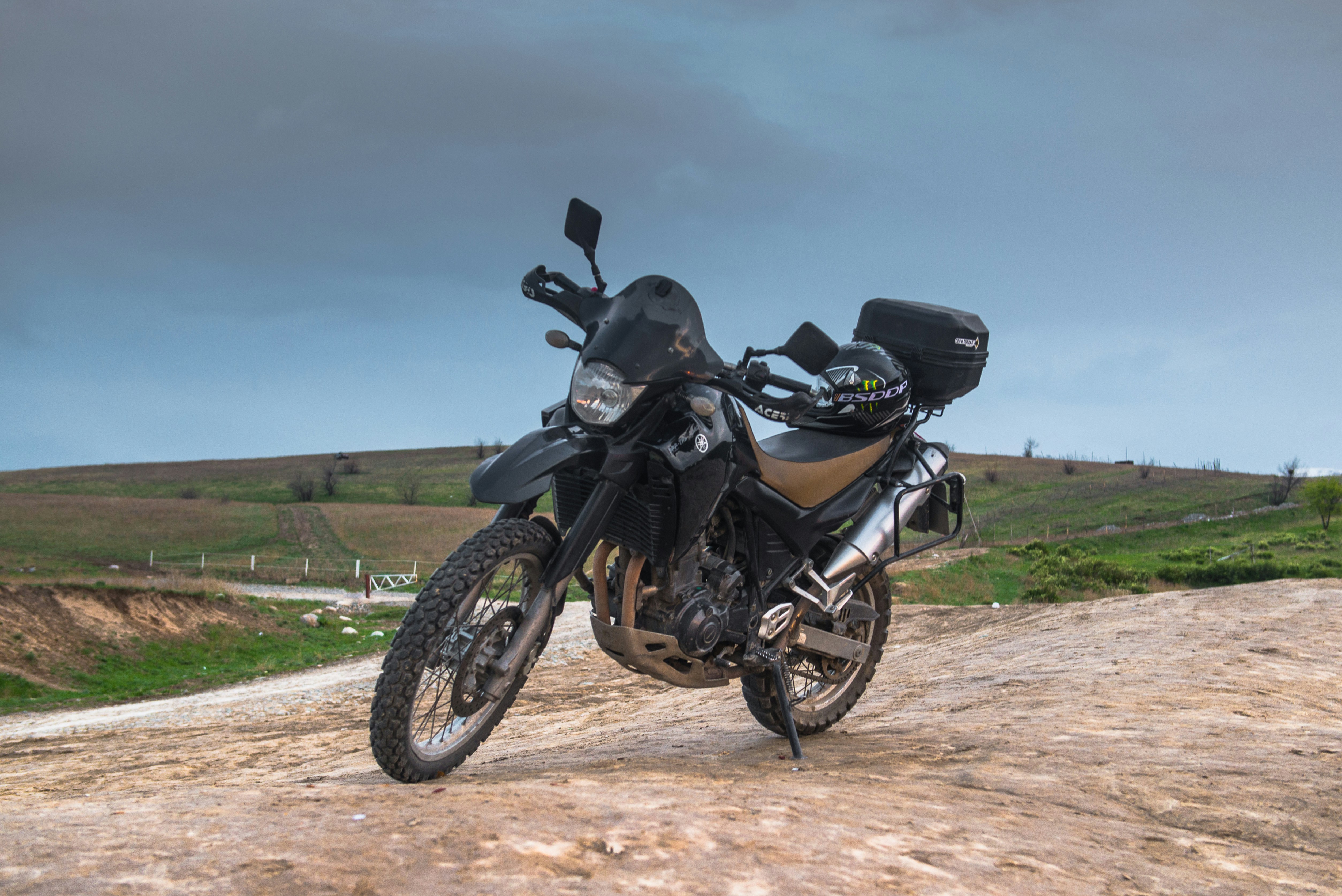Adventure motorcycle on rugged mountain trail, dust clouds, wide handlebars, knobby tires, dramatic highland landscape, overcast sky, powerful stance