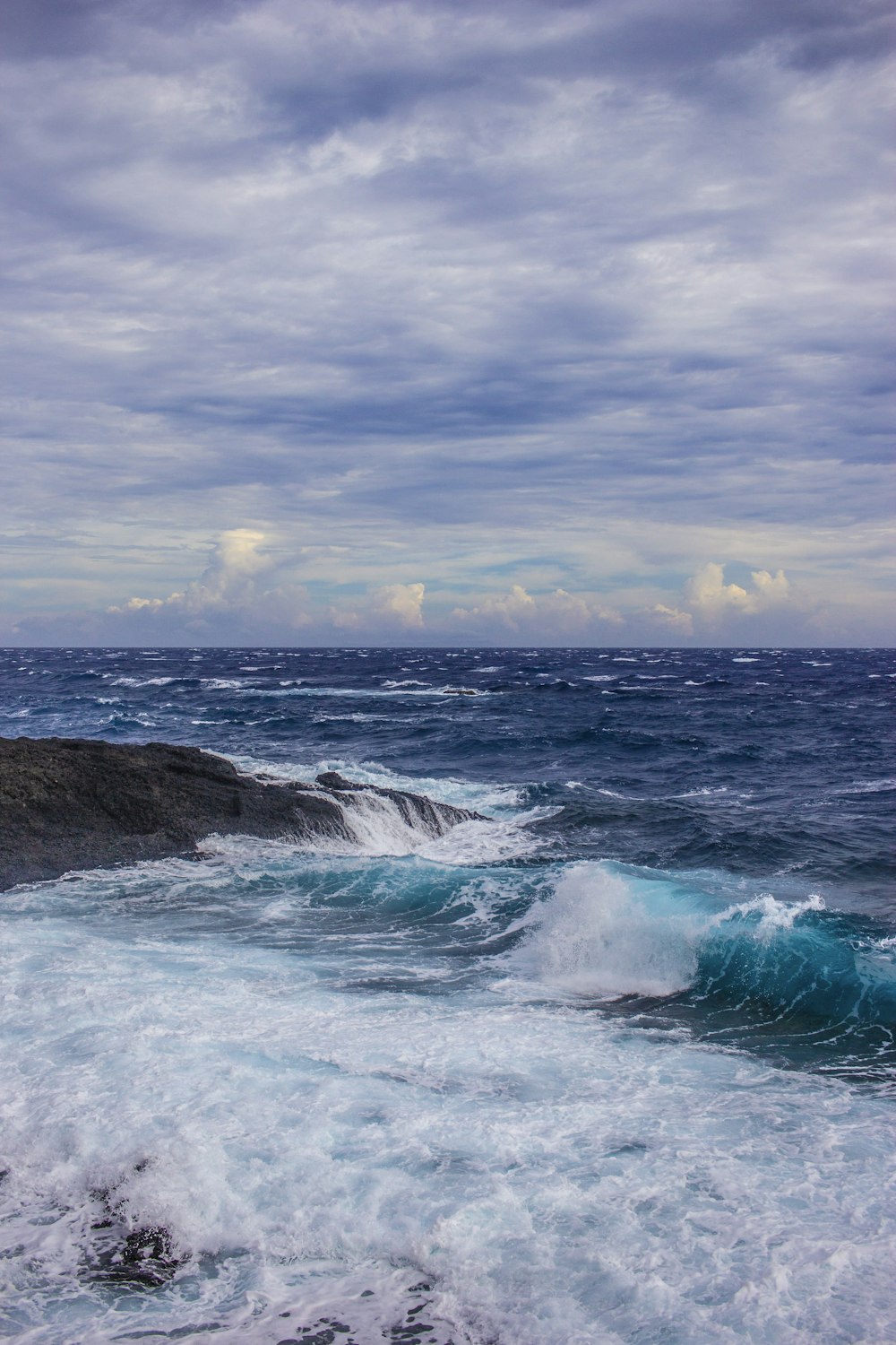 ondas do oceano quebrando em formação rochosa negra sob nuvens brancas durante o dia