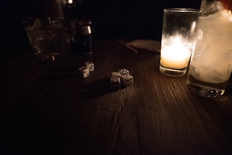 A dimly lit scene featuring a wooden table with several dice scattered across it. Two glasses are visible, one containing a candle that provides soft lighting, while the other appears to hold a drink with ice cubes. A lighter is also seen on the table.
