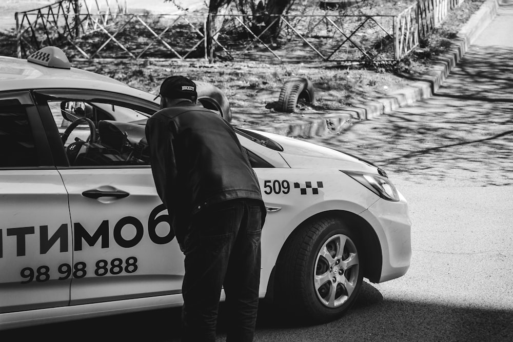 grayscale photo of man in black jacket standing beside white car