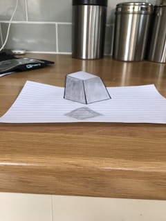A piece of lined paper placed on a wooden surface with a realistic 3D pencil drawing of a geometric shape, creating an optical illusion. The drawing appears to be hovering above the paper. In the background, there are large metallic kitchen canisters and a countertop with a tiled backsplash.