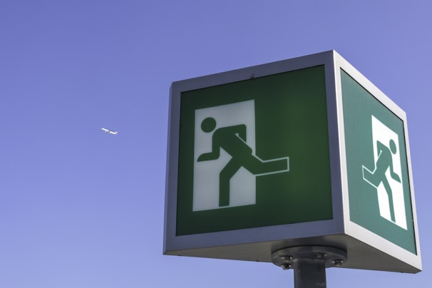 A large, green emergency exit sign displayed on a post against a clear blue sky. The sign features a white figure in motion with an arrow, indicating the direction towards the exit. In the background, a small airplane is visible, flying high in the sky.
