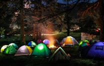 Multiple colorful tents are pitched in a forested area at night, surrounding a glowing campfire. The trees are tall and dense, creating a canopy above. Soft light from the fire illuminates the tents, creating a cozy and communal atmosphere. Shadows play across the forest floor, and a hint of moonlight or distant lights can be seen through the trees.