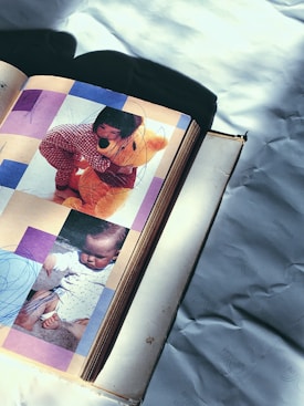A vintage photo album is open to a page displaying two childhood photographs. The top image shows a young child in red checkered clothing hugging a large teddy bear. The bottom image features a baby dressed in white sitting outdoors. The photographs are overlaid with scribbles, and the album is lying on a surface with a shadow cast over part of it.