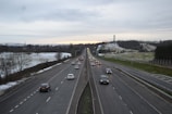 A highway stretches into the distance with multiple lanes of cars traveling in both directions. The road is flanked by fields that are lightly dusted with snow. On the right side, a tall tower is visible on a hill. The sky is overcast, and the landscape is peaceful with a hint of the countryside visible in the background.