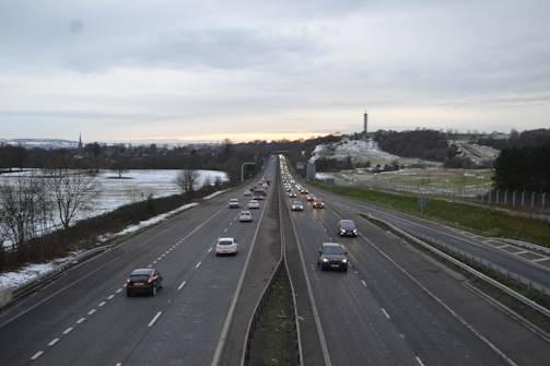 A highway stretches into the distance with multiple lanes of cars traveling in both directions. The road is flanked by fields that are lightly dusted with snow. On the right side, a tall tower is visible on a hill. The sky is overcast, and the landscape is peaceful with a hint of the countryside visible in the background.