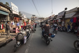 A busy street market scene with various shops displaying colorful items on both sides. People are walking and riding scooters through the narrow street. Goods such as clothing and decorations are visible, with signs and banners in a local language above the shops.