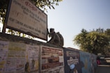 A monkey sits atop a wall covered with various advertisements and posters. Nearby, there is a sign displaying information about a location in both Hindi and English. Trees with green leaves are visible in the background, and a clear blue sky is overhead.