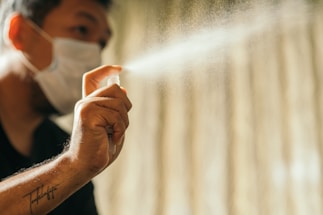 A person wearing a mask sprays a fine mist from a bottle. The background is softly blurred, drawing attention to the hand and the spray.