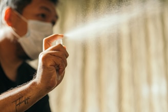 A person wearing a mask sprays a fine mist from a bottle. The background is softly blurred, drawing attention to the hand and the spray.