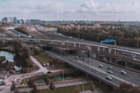 Aerial view of a complex highway and railway interchange surrounded by green trees and a partly cloudy sky. Cars and trucks can be seen traveling on the different layers of roads while trains pass by on elevated tracks. The urban skyline is visible in the distance.