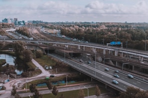 Aerial view of a complex highway and railway interchange surrounded by green trees and a partly cloudy sky. Cars and trucks can be seen traveling on the different layers of roads while trains pass by on elevated tracks. The urban skyline is visible in the distance.
