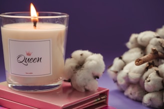 A lit candle in a glass holder labeled 'Queen' is placed on top of two stacked pink notebooks. Cotton branches are positioned to the right of the candle. The background is a deep purple, creating a cozy and serene atmosphere.