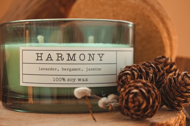A green-glass jar candle labeled 'Harmony' with scents of lavender, bergamot, and jasmine, made from 100% soy wax. Pine cones are artistically arranged next to the candle, creating a rustic and cozy setting.