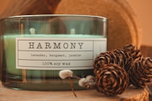 A green-glass jar candle labeled 'Harmony' with scents of lavender, bergamot, and jasmine, made from 100% soy wax. Pine cones are artistically arranged next to the candle, creating a rustic and cozy setting.