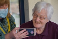 An older woman with white hair and glasses smiles while looking at a smartphone being held by another person wearing a face mask. The person holding the phone has shoulder-length hair and a scarf with yellow and gray patterns.