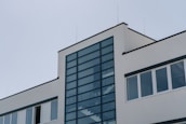 A modern building with a minimalist architectural design, featuring a large glass panel section in the center framed by dark metal. The structure has white walls and several rectangular windows.