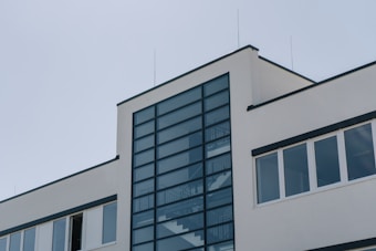 A modern building with a minimalist architectural design, featuring a large glass panel section in the center framed by dark metal. The structure has white walls and several rectangular windows.