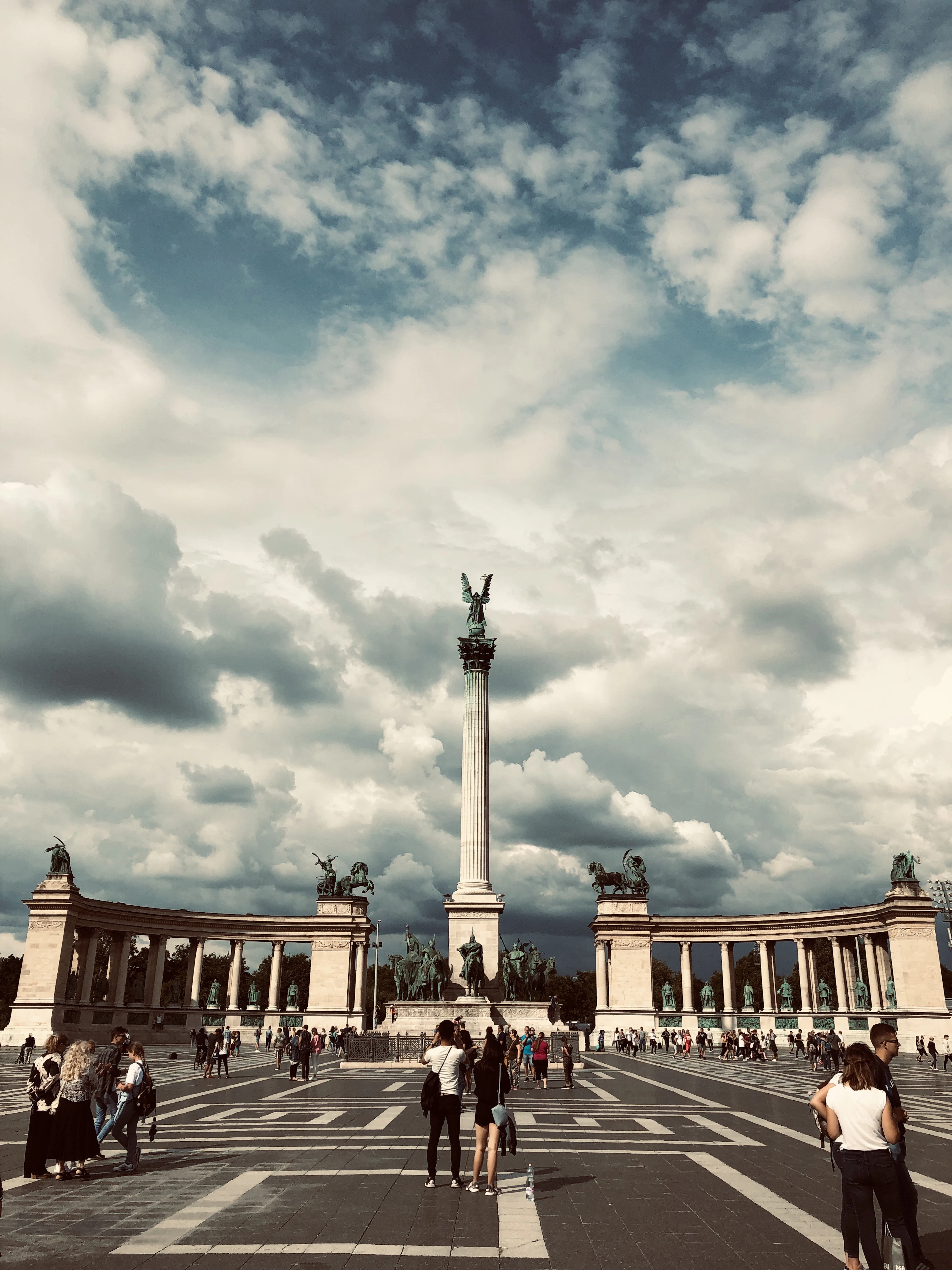 Heroes' Square in Budapest beneath a towering bank of storm cloud, the Millennium Monument column and its curved colonnades of bronze statues rising above visitors scattered across the patterned stone paving
