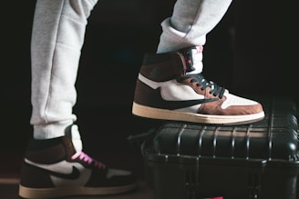 A pair of stylish high-top sneakers with brown, black, and white color combinations on different surfaces stands prominently. One shoe rests on a sturdy black case while the person is wearing light gray sweatpants.