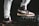 A pair of stylish high-top sneakers with brown, black, and white color combinations on different surfaces stands prominently. One shoe rests on a sturdy black case while the person is wearing light gray sweatpants.